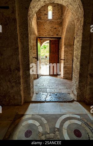 Ein historischer Steinbogen im Kloster Andromonastiro in Messinia, Griechenland, mit einer offenen Holztür, die zu einem sonnendurchfluteten Innenhof führt. Stockfoto