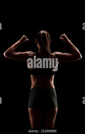 Ein kraftvolles, hintergrundbeleuchtetes Bild einer Frau in Fitnesskleidung, das Stärke und Entschlossenheit zeigt, indem sie ihre Armmuskeln vor schwarzem Hintergrund beugt. Stockfoto