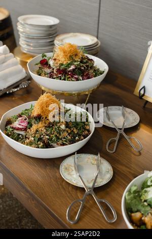 Große Schüsseln mit bunten Salaten gefüllt mit Gemüse und Belägen sind auf einem Holztisch an einer Buffetstation angeordnet. Teller und Servierutensilien sind vorhanden Stockfoto