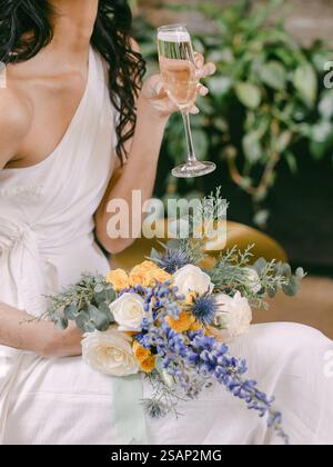 Eine Frau in einem fließenden weißen Kleid hält ein Glas Champagner, während sie anmutig sitzt. Sie präsentiert einen wunderschönen Blumenstrauß mit Rosen und Lavendel A Stockfoto