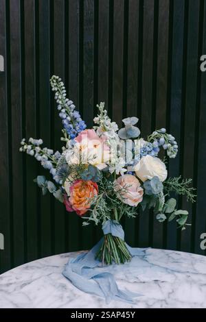 Elegantes Bouquet mit pastellfarbenen Rosen, blauen Blumen und Eukalyptus, wunderschön auf einem Marmortisch vor dunkler Holzkulisse angeordnet Stockfoto