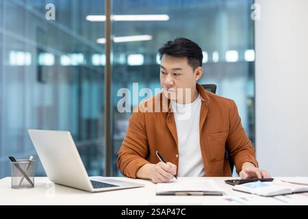 Seriöser, zuversichtlicher asiatischer Finanzier, der im Büro an Papierkram arbeitet, Geschäftsmann mit Taschenrechner, der Finanzberichte und Steuerformulare im Büro mit Laptop ausfüllt. Stockfoto