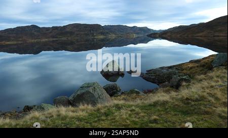 Ruhige Landschaft mit ruhigem Wasser, das tagsüber den bewölkten Himmel auf einem See vom felsigen Ufer reflektiert. Norwegen Stockfoto