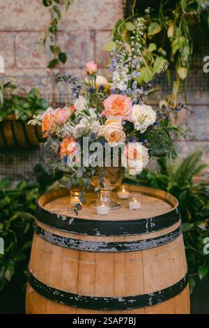 Ein wunderschön gestalteter Blumenstrauß mit leuchtenden Rosen und Grün sitzt auf einem Holzfass, umgeben von üppigen Pflanzen. Mit dieser Einstellung wird ein warmes Bild aufgenommen Stockfoto
