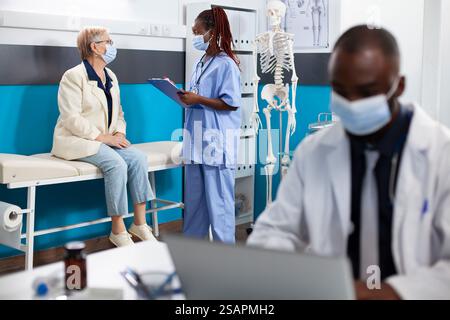 afroamerikanische Krankenschwester hält Klemmbrett und fragt kaukasische alte Frau über ihre Krankengeschichte als Arzt auf Laptop in der Nähe. Schwarze Frau in blauen Schutteln, die mit älteren Patienten spricht. Stockfoto