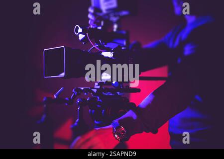 Ein Kamerabediener passt die Filmausrüstung sorgfältig mit farbenfroher Beleuchtung im Hintergrund an, um die perfekte Aufnahme zu erzielen. Stockfoto