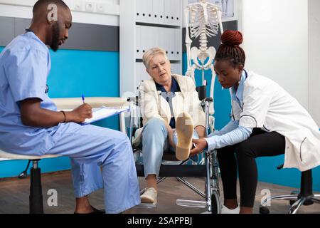 Der Arzt hilft alte Frau im Rollstuhl während eines Arzttermins, während die männliche Krankenschwester Notizen auf dem Klemmbrett macht. Aufmerksames medizinisches Personal bietet eine personalisierte Betreuung für pensionierte Patienten, die Hilfe benötigen. Stockfoto