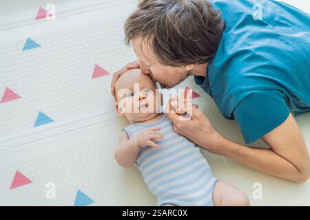 Ein Baby liegt auf einer weichen Kinderspielmatte, während seine Vaterhände sie sanft stützen und schützen. Herzerwärmender Moment der Pflege und Verbindung Stockfoto