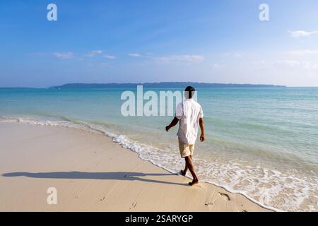 Isolierter Mann, der am unberührten weißen Sandstrand mit sanften Meereswellen am Morgen aus einem flachen Winkel spaziert, wird am Radhanagar Beach auf der Havelock Island aufgenommen Stockfoto