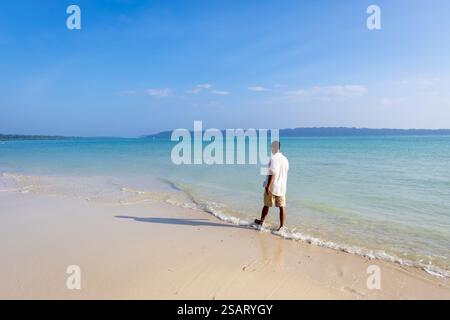 Isolierter Mann, der am unberührten weißen Sandstrand mit sanften Meereswellen am Morgen aus einem flachen Winkel spaziert, wird am Radhanagar Beach auf der Havelock Island aufgenommen Stockfoto