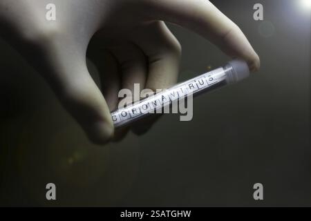 Hand in einem Handschuh, der ein mit Coronavirus markiertes Reagenzglas in schwachem Licht hält Stockfoto