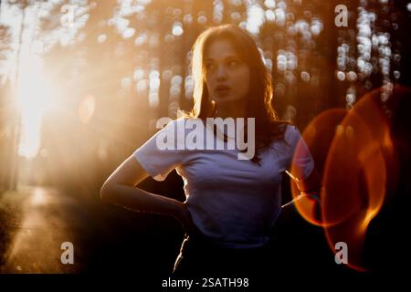 Eine Frau in einem weißen Hemd, von der Sonne beleuchtet, steht in einem Wald mit nachdenklichem Ausdruck Stockfoto