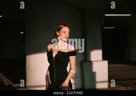 Eine Frau in einem eleganten schwarzen Kleid steht selbstbewusst im Sonnenlicht mit urbanen Schatten Stockfoto