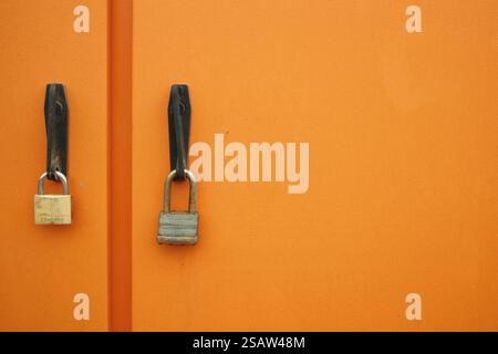 Zwei Vorhängeschlösser an einer orangefarbenen Metalltür symbolisieren Sicherheit, Schutz und Zugangskontrolle. Stockfoto