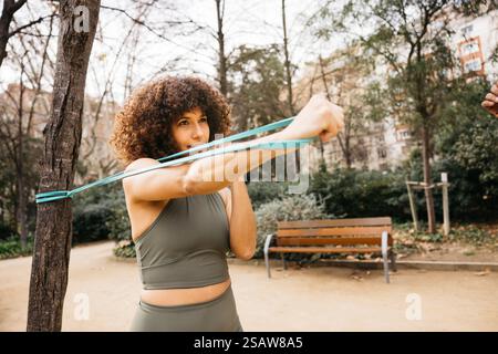 Eine Frau in Sportbekleidung führt Krafttraining mit einem elastischen Widerstandsband in einer von Grün und Bänken umgebenen Parklandschaft durch. Stockfoto