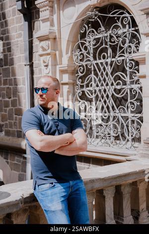 Mann in Brille gefaltete Hände auf Brust vor dem Schloss, gekleidet in Jeans, weiße Turnschuhe und ein blaues Poloshirt, eine mittelalterliche Burg aus Stockfoto