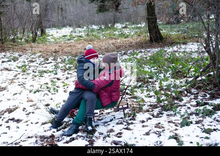 Eltern und Kind teilen einen zärtlichen Moment auf einem Schlitten inmitten eines leicht verschneiten Waldes. Das Kind liegt in der Nähe und genießt das Winterwunderland. Stockfoto