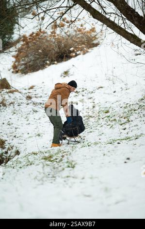 Ein Mann in brauner Jacke schiebt ein Kind sanft auf einem kleinen Schlitten einen schneebedeckten Hügel hinunter. Leichter Schnee fällt, und die Szene ist friedlich und fröhlich. Stockfoto