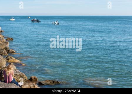 Eine Frau mit langen blonden Haaren sieht man von hinten auf einem Felssteg mit Blick aufs Meer, wo Leute auf Booten fischen, Ponce Inlet, Florida. Stockfoto