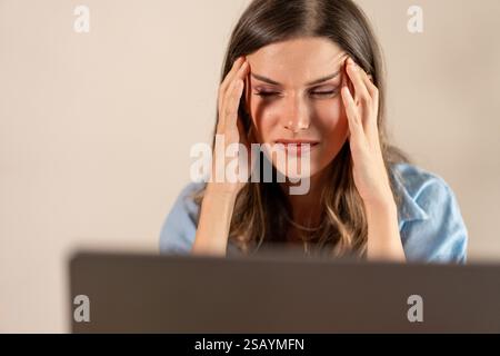 Eine frustrierte Frau, die den Kopf hält, während sie an einem Laptop arbeitet, und die in einer Home Office-Umgebung unter Stress und Kopfschmerzen leidet. Stockfoto