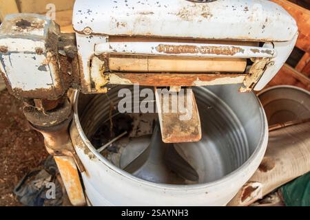 Eine alte Waschmaschine mit Holzgriff sitzt in einem schmutzigen Hof. Die Maschine ist rostig und hat ein abgenutztes Aussehen Stockfoto