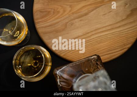 Blick von oben auf ein Whiskey-Glas und eine Karaffe auf einer schwarzen Oberfläche mit einem Holzbrett, die eine warme, elegante Atmosphäre schaffen. Kopierraum für Text. Stockfoto
