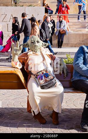 Straßenkünstler verkleidet als Yoda, der sich während des Valletta-Karnevals auf Malta auf einer Bank im belebten öffentlichen Raum entspannt Stockfoto