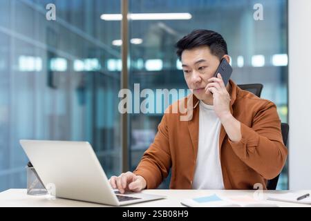 Seriöser und zielgerichteter erfolgreicher asiatischer Geschäftsmann, der über Mobiltelefon spricht. Männlicher Büroangestellter am Arbeitsplatz im Büro mit Laptop. Stockfoto