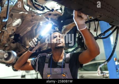Junger Mechaniker mit Taschenlampe, der die Unterseite des auf dem Aufzug angehobenen Fahrzeugs in einer professionellen Garage überprüft Stockfoto