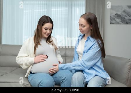 Lächelnde schwangere Frau im dritten Trimenon, die auf dem Sofa sitzt, während ihre Freundin ihren Bauch vorsichtig berührt. Mutterschaftsbegriff, Freundschaft Stockfoto