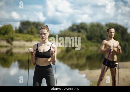Frau und Mann, die sich auf Widerstandsbandübungen an einem See mit Bäumen im Hintergrund konzentrieren Stockfoto