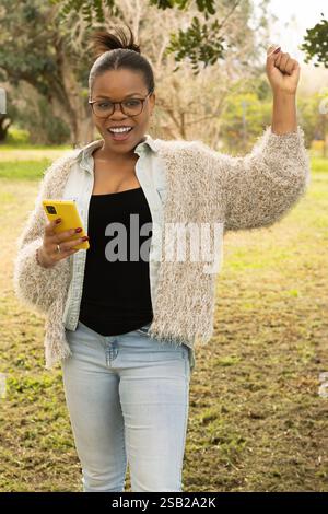 Eine fröhliche schwarze Frau, die eine Brille und ein gemütliches Outfit trägt, feiert, während sie ihr Smartphone ansieht. Stockfoto