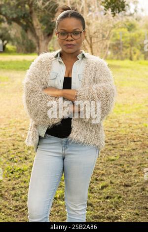 Eine schwarze Frau mit Brille und gemütlichem Outfit steht mit überkreuzten Armen und zeigt einen ernsten und entschlossenen Ausdruck. Stockfoto