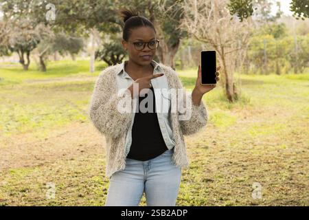 Eine selbstbewusste schwarze Frau steht draußen und lächelt, während sie mit einem leeren Bildschirm auf ein Smartphone zeigt. Stockfoto
