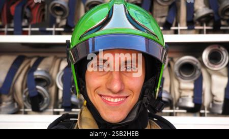 Junger, lächelnder Feuerwehrmann, der nach dem Feuer in voller Ausrüstung vor dem Hintergrund eines großen roten Trucks in die Kamera schaut. Porträt eines glücklichen männlichen Feuerwehrmannes Stockfoto