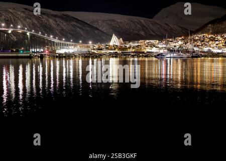Nachtblick auf Tromso, Norwegen. Beleuchtete Gebäude, die Arktische Kathedrale und Reflexionen auf dem Wasser, vor dem Hintergrund schneebedeckter Berge Stockfoto