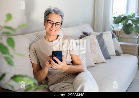 Eine ältere Frau mit grauem Haar sitzt auf der Couch, lächelt und posiert für die Kamera. Stockfoto