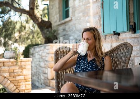 Eine Frau entspannt sich auf einer Terrasse, schlürft Kaffee im warmen Sonnenlicht, umgeben von rustikalen Steinmauern und üppigem Grün, und genießt einen ruhigen Start in die Stockfoto