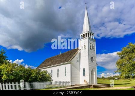 Eine weiße Kirche mit einem Turm und einem Hund davor. Der Himmel ist bewölkt und die Sonne scheint Stockfoto