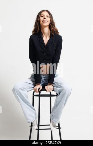 Eine modische Frau sitzt auf einem schwarzen Hocker, trägt ein schwarzes Oberteil und Jeans und posiert selbstbewusst vor einem schlichten Hintergrund in einem gut beleuchteten modernen Studio. Stockfoto