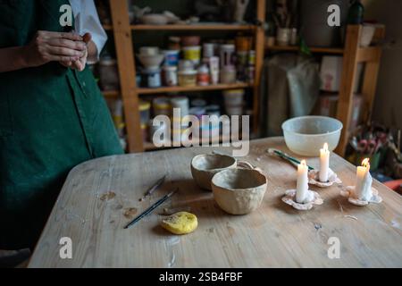 Handwerker arbeiten an einem Holztisch in der Töpferwerkstatt mit Schnitzwerkzeugen, Schwamm und weißen Tonbechern Stockfoto