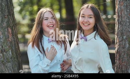 Zwei fröhliche Schulmädchen in lila Fliege lachen in einem sonnigen Park, der von Bäumen umgeben ist Stockfoto