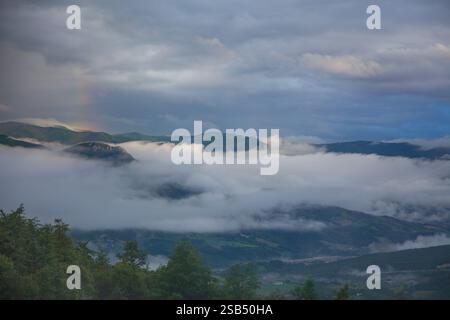 Italien, Apennin. Abendlicher bewölkter Himmel über einem grünen Bergtal. Geringer dicker Nebel und ein Stück Regenbogen Stockfoto
