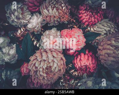 Ein üppiges Arrangement künstlicher Blumen in gedämpften Rosa-, Burgunderrot- und Cremetönen. Dunkelgrüne Blätter und goldfarbene Akzente verleihen Tiefe. Vintage-Ästhetik. Stockfoto