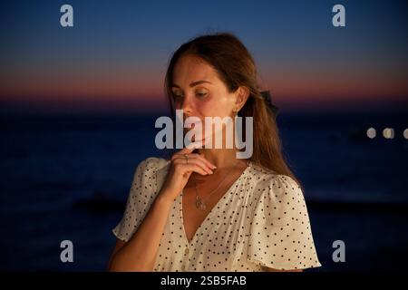 Nachdenkliche Frau im Tupfen-Kleid bei Sonnenuntergang am Meer Stockfoto