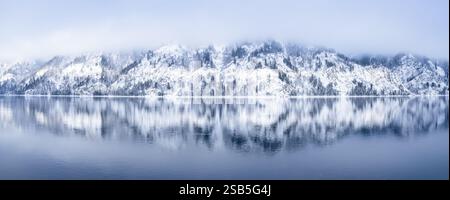 Eine friedliche und ruhige Winterszene mit atemberaubenden schneebedeckten Gipfeln und einem spiegelähnlichen See unter sanften Wolken, schneebedeckten Bergen mit Kiefern Stockfoto