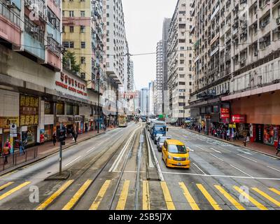 Hongkong, China - 01. Februar 2025 : Eine Straßenszene in Hongkong mit einer Straßenbahnstrecke, die von hohen Gebäuden und geschäftigen Geschäften flankiert wird. Fußgänger und Stockfoto