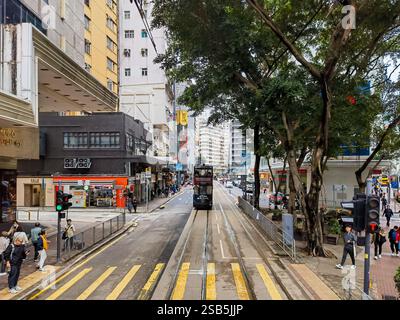 Hongkong, China - 01. Februar 2025 : Eine Straßenbahn aus Hongkong gleitet über eine Stadtstraße, flankiert von hohen Gebäuden und üppigen grünen Bäumen. Fußgänger spazieren Stockfoto