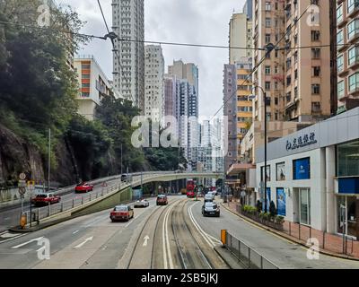 Hongkong, China - 01. Februar 2025 : Eine Straßenbahn aus Hongkong gleitet entlang einer Stadtstraße, flankiert von hohen Gebäuden und üppigem Grün. Die Fahrbahnkurven reichen Stockfoto