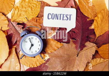 Eine Uhr sitzt auf einem Blätterhaufen, in der das Wort Contest in einer Sprechblase geschrieben ist, was Wettbewerb, Termine und zeitabhängiges chall symbolisiert Stockfoto
